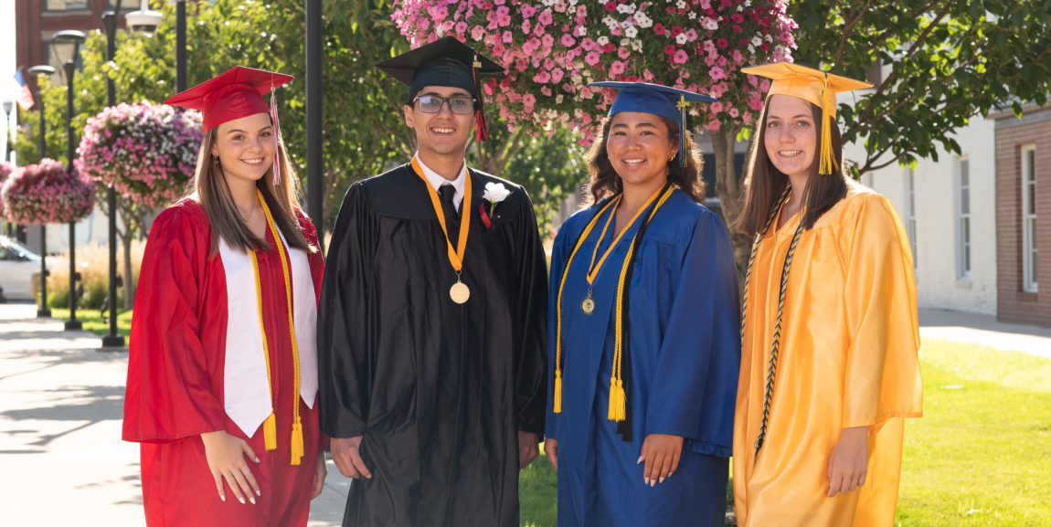 Four students standing in various colors of caps and gowns. In the background is a plaza area with sidewalks, grass, trees and hanging flower baskets.