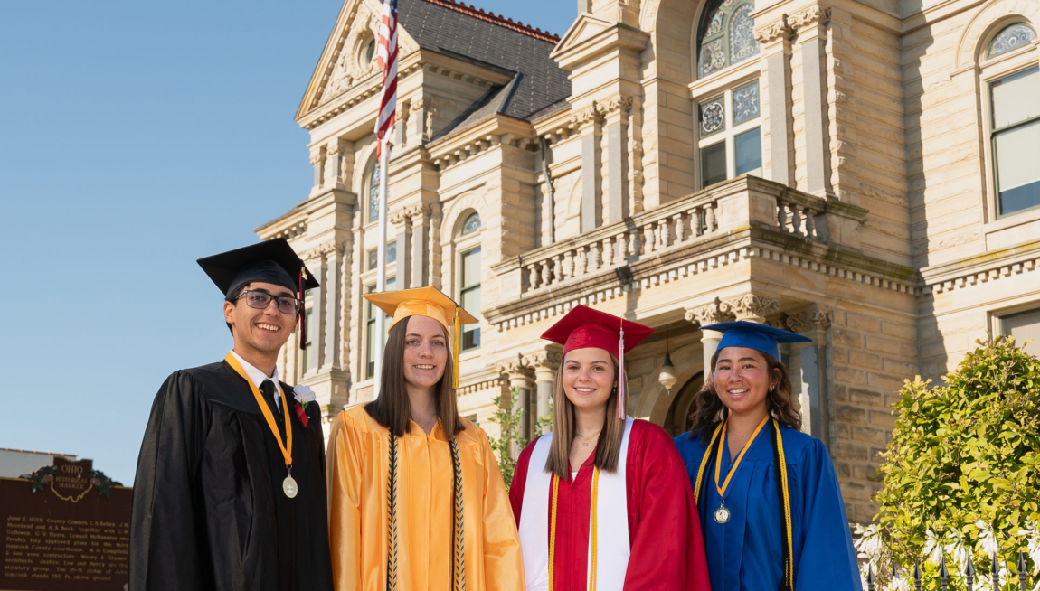 Four students in various colors of caps and gowns pose together in front of the  Hancock County Courthouse.